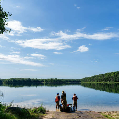Three individuals stand at the edge of a calm lake under a bright blue sky, surrounded by lush green trees and vegetation.